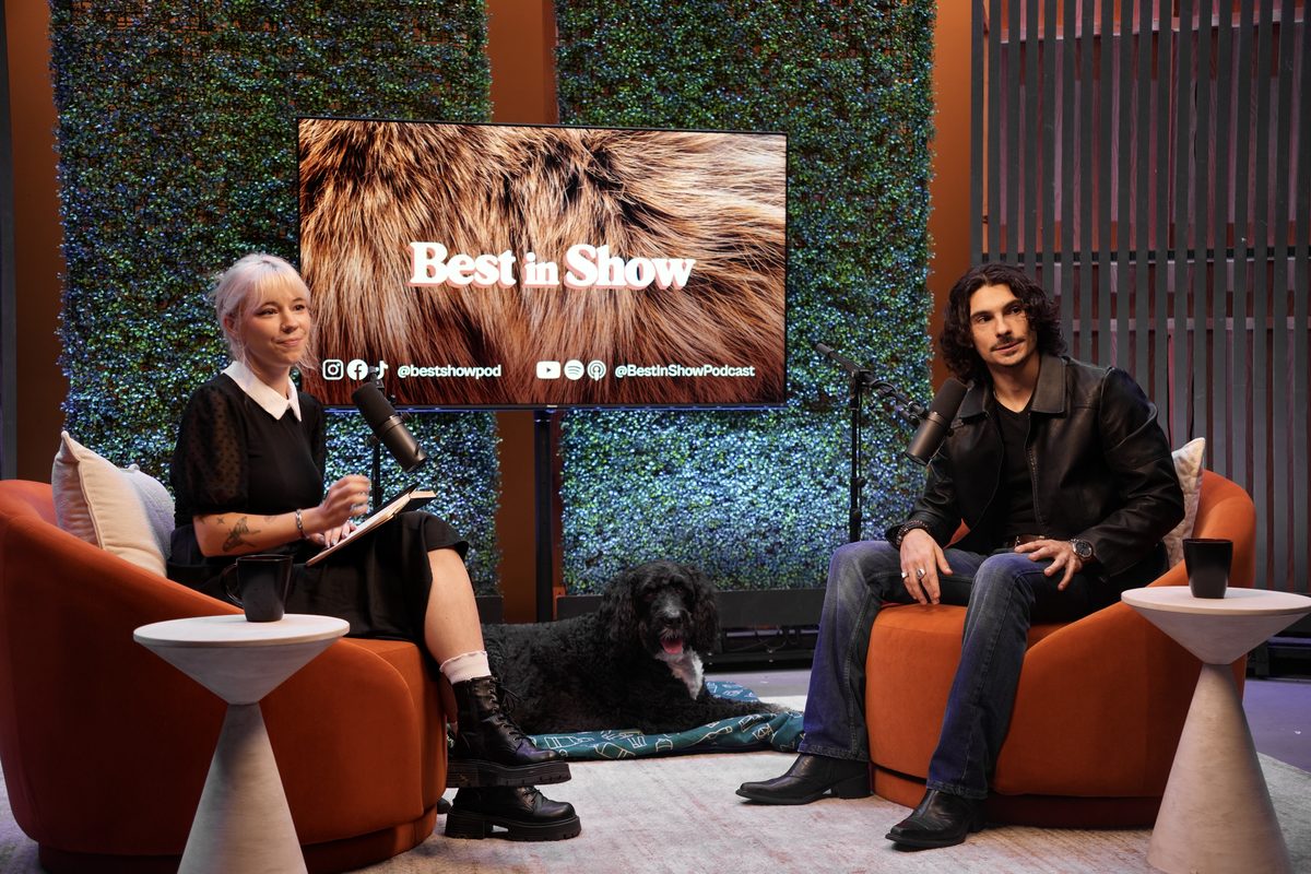 Mimi interviewing a guest on set, a black dog resting at the foot of his chair